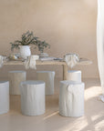 Hydro-dipped concrete dining table styled with white timber stump stools, neutral linen napkins, and ceramic tableware. A soft, sunlit setting with organic textures and natural elements, creating a relaxed, earthy ambiance