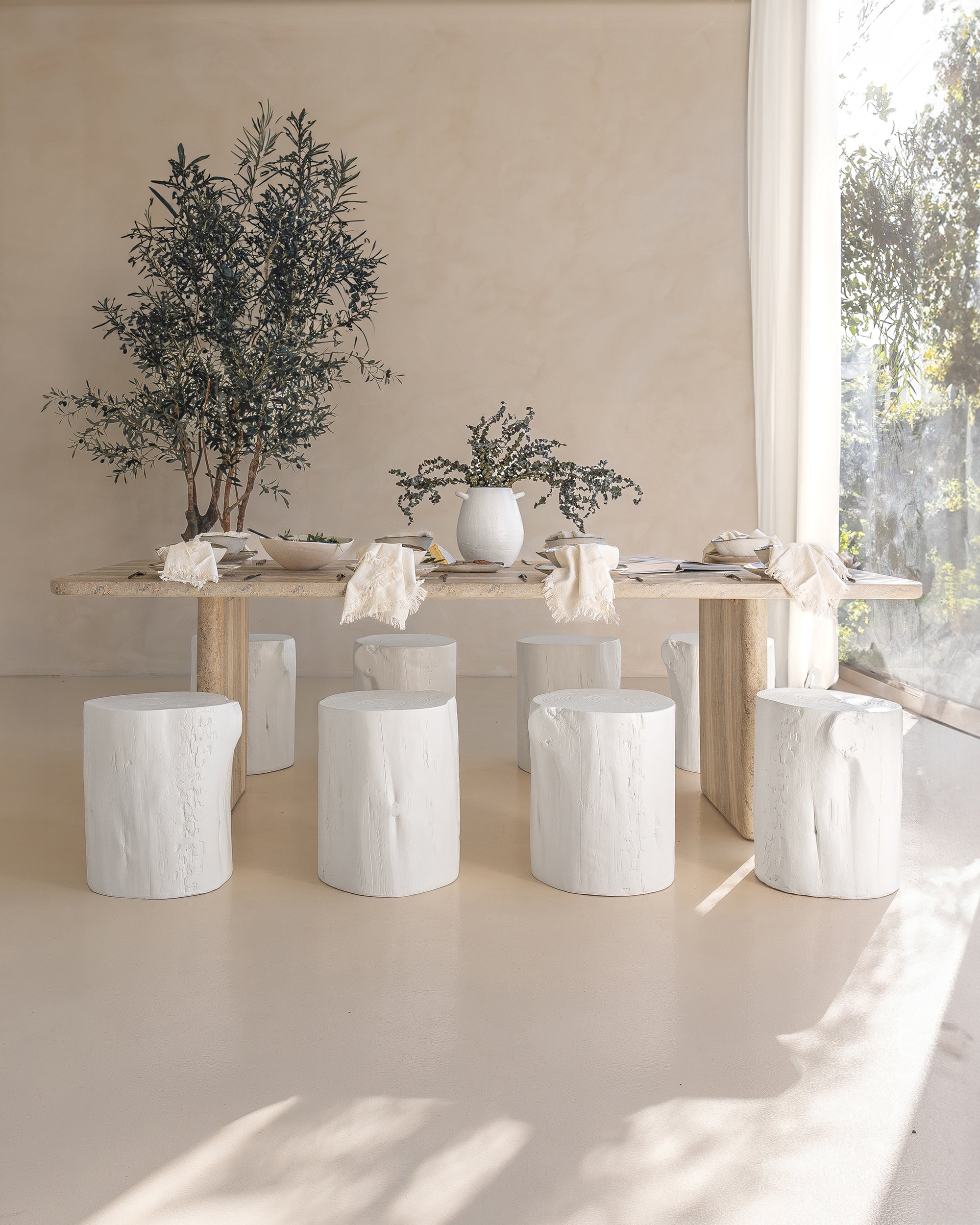 Hydro-dipped concrete dining table styled with white timber stump stools, neutral linen napkins, and ceramic tableware. A soft, sunlit setting with organic textures and natural elements, creating a relaxed, earthy ambiance