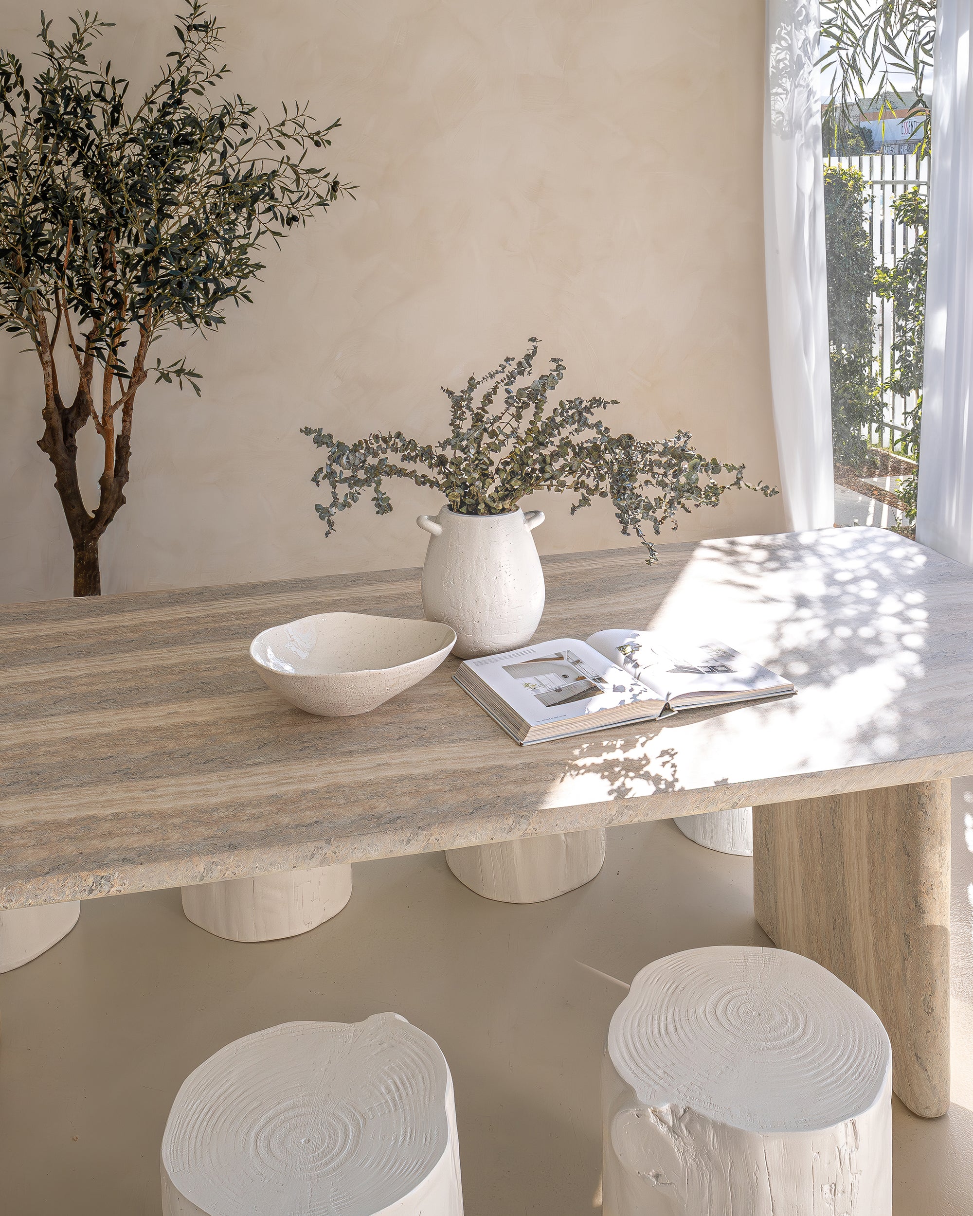 Hydro-dipped concrete dining table styled with white timber stump stools, neutral linen napkins, and ceramic tableware. A soft, sunlit setting with organic textures and natural elements, creating a relaxed, earthy ambiance