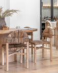 Close-up of natural fibre dining chairs with curved backrests and timber frames around a round timber table in a light-filled kitchen