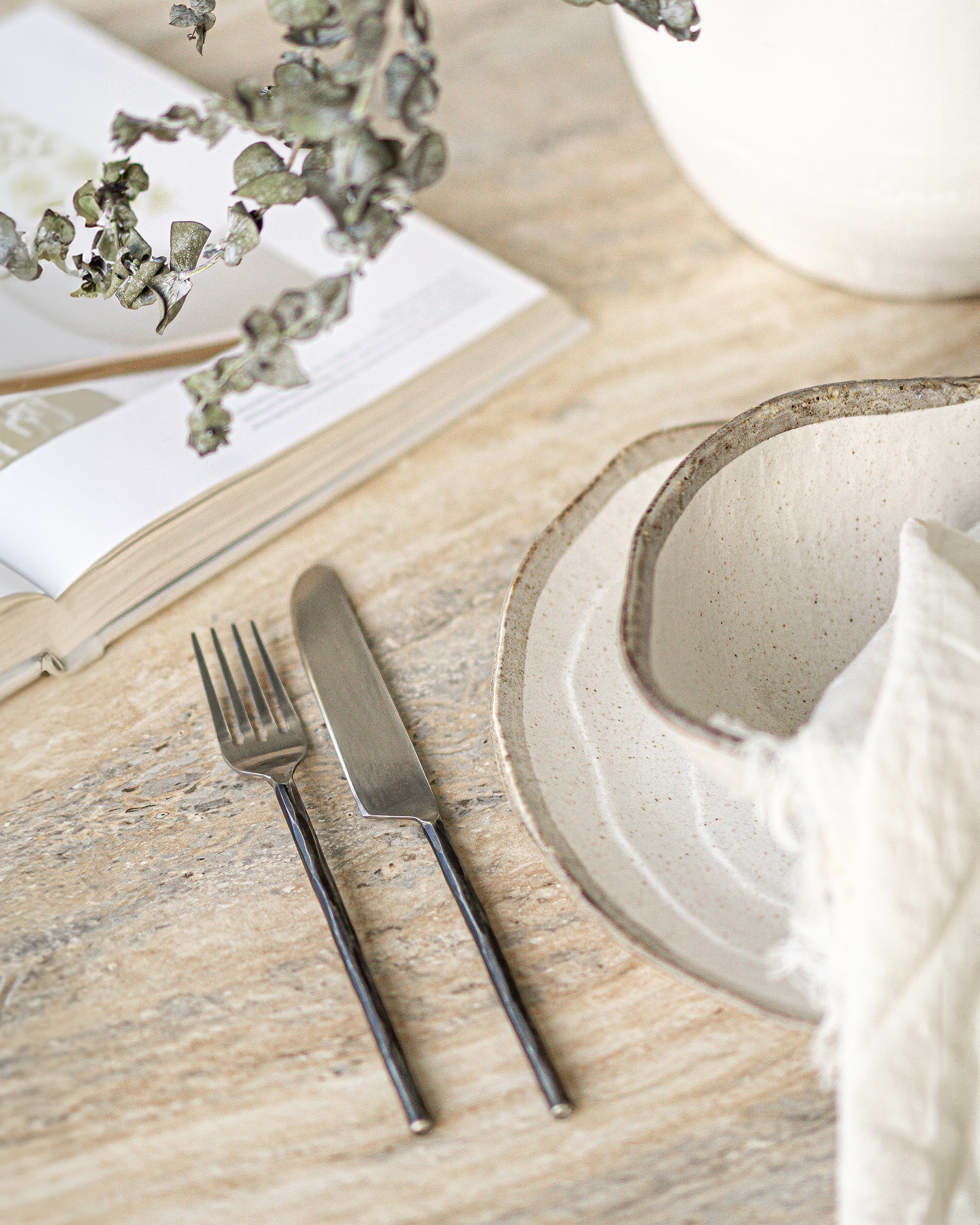 A minimalist table setting featuring hand-forged stainless steel cutlery with sleek black handles, paired with rustic ceramic dishware on a textured stone table, evoking a refined yet organic dining experience.