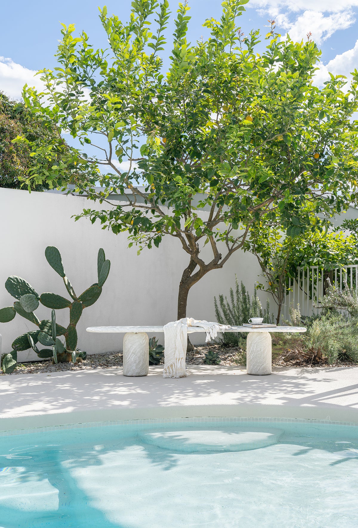 KULALA Living Ayanda Concrete Bench in Eggshell styled with the Fara Linen Bath Towel and Small Throw beside a swimming pool, surrounded by a leafy citrus tree, cacti and lush coastal garden greenery.