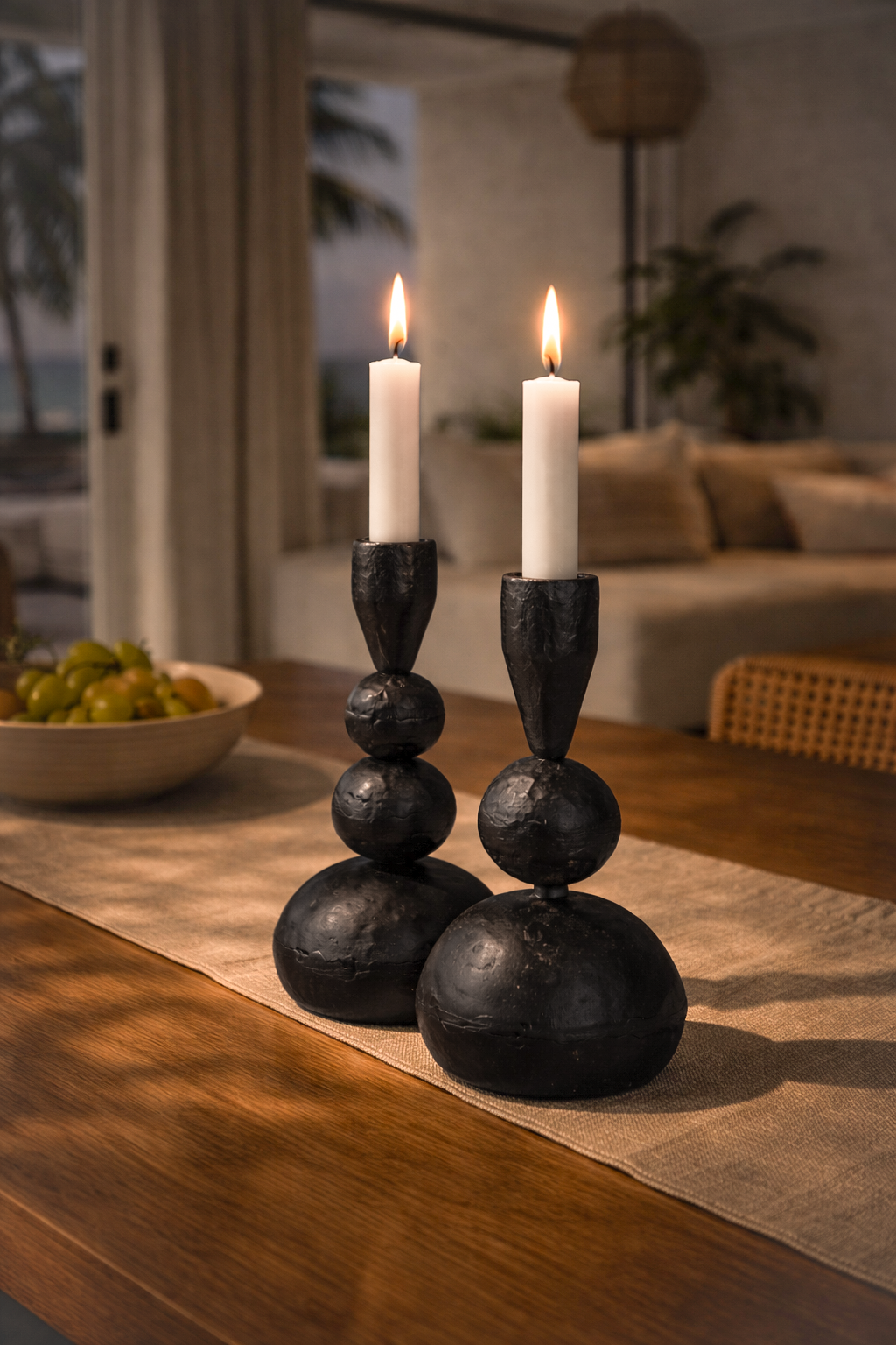 Black ball candle holders styled on a timber dining table with lit white taper candles, soft linen runner and warm ambient evening light.