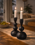 Black ball candle holders styled on a timber dining table with lit white taper candles, soft linen runner and warm ambient evening light.