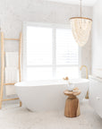 A serene bathroom featuring a freestanding white bathtub, a natural wooden ladder towel rack, and a sculptural wooden side table. Accented with a cascading shell chandelier and brass tapware, creating a tranquil spa-like ambiance
