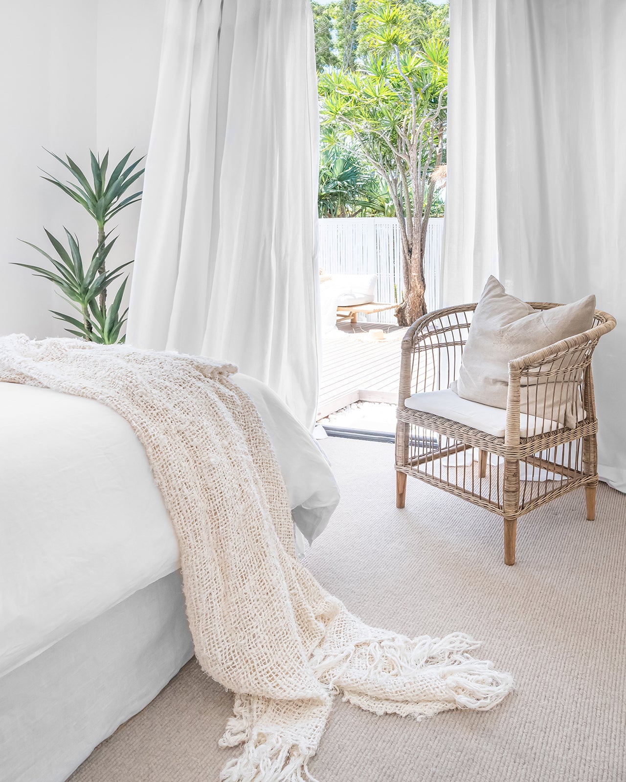 A serene bedroom featuring a neutral-toned upholstered bed with soft layered bedding, a natural rattan armchair with textured cushion for a calming retreat.