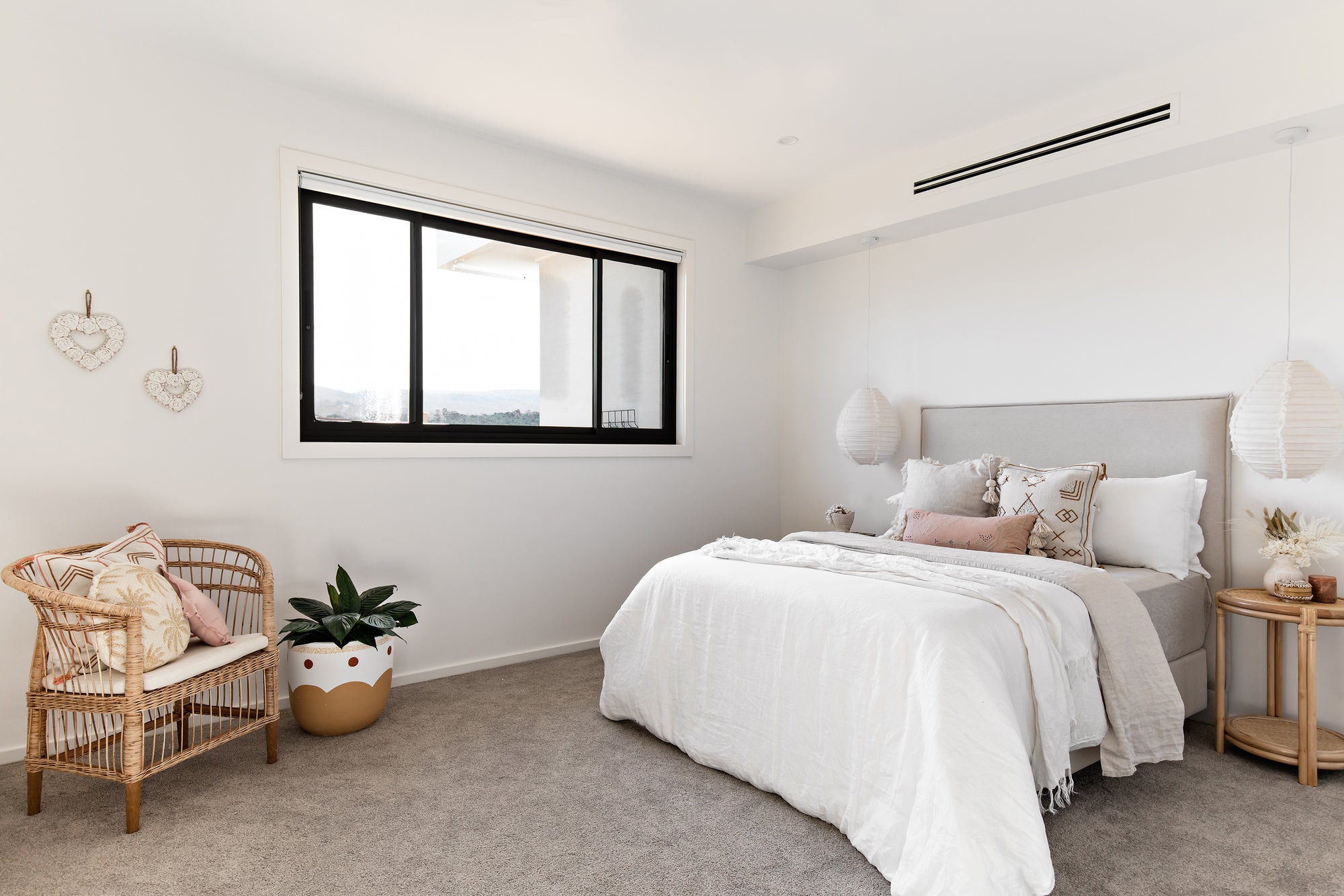 A serene bedroom featuring a neutral-toned upholstered bed with soft layered bedding, a natural rattan armchair with textured cushions, and delicate pendant lighting for a calming retreat.