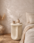 Berber side table in driftstone finish with sculptural curved form styled beside a bed in a soft neutral bedroom setting.