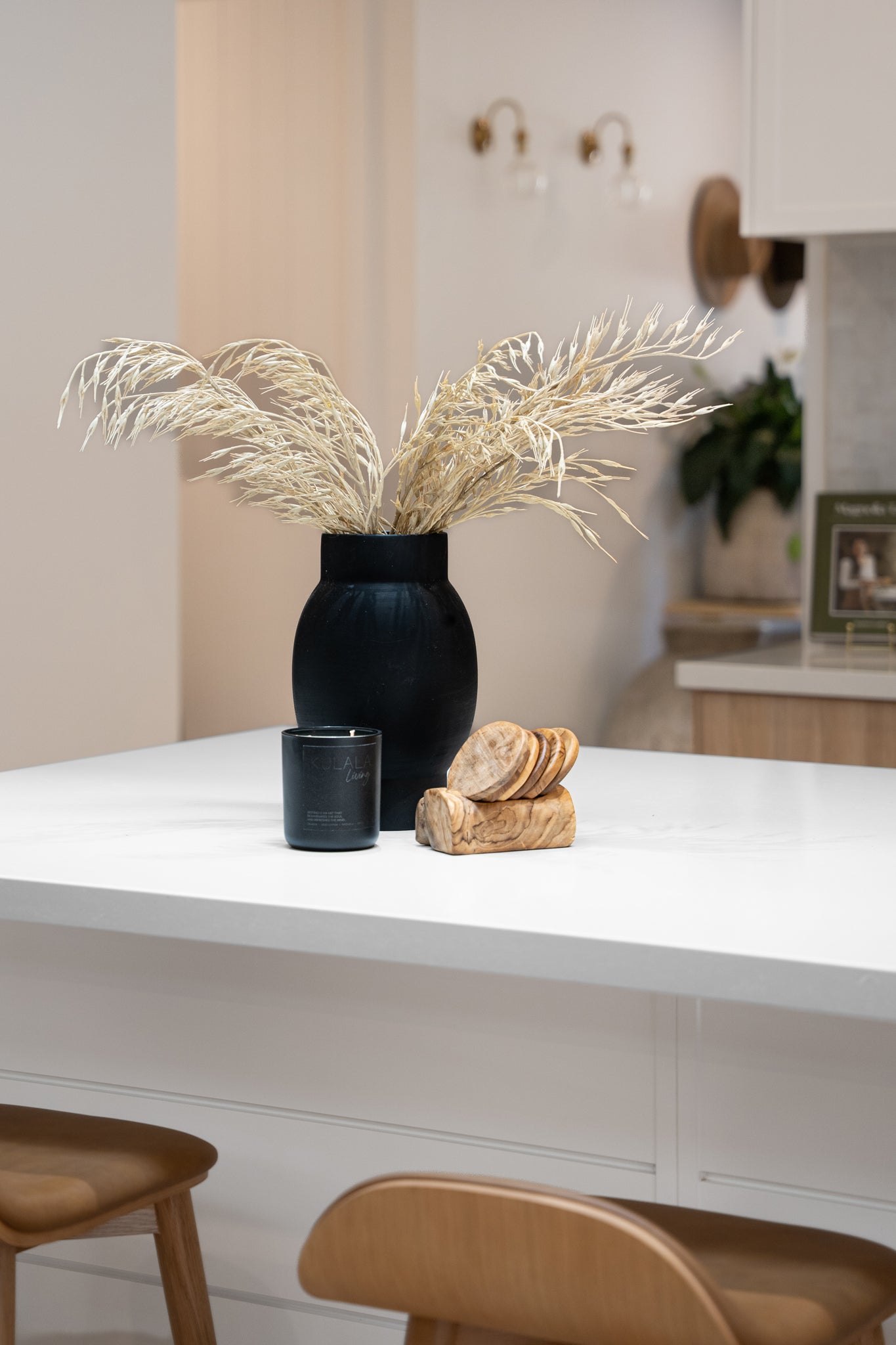 A modern kitchen countertop featuring a striking black ceramic vase with dried foliage, a black Kulala Living candle, and a wooden coaster set. The scene is complemented by warm wood tones and a neutral palette, creating a refined and inviting aesthetic