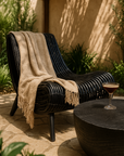Black rattan outdoor Congo lounger styled with a beige throw in a shaded patio setting beside a round black timber table with an espresso martini.