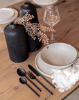 A contemporary dining table setting featuring KULALA Living’s sleek black cutlery, speckled ceramic plates, and a soft linen napkin on a warm timber table. Styled with black sculptural vases and delicate dried foliage, this setting embodies modern elegance and refined simplicity