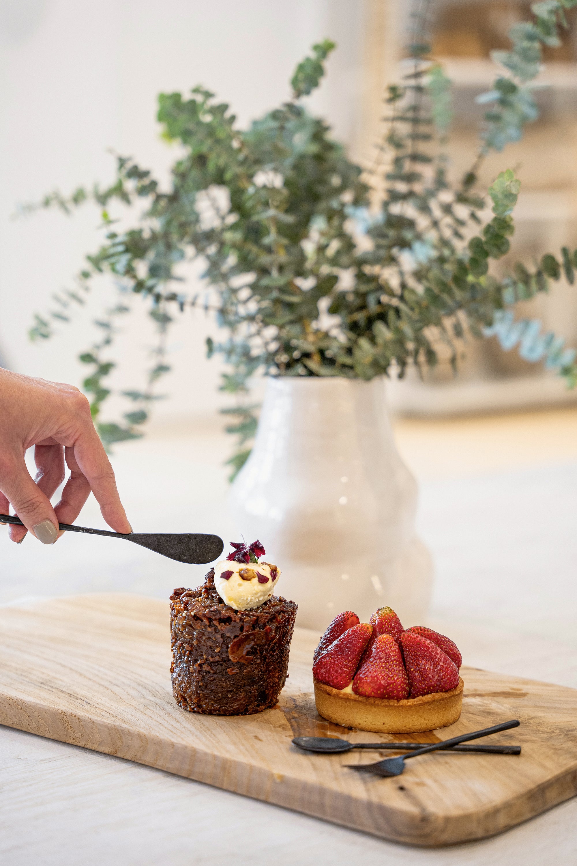 A delicate dessert moment featuring KULALA Living’s black hand-forged butter knife, elegantly spreading cream onto a caramelised fig and nut pastry. Paired with a fresh strawberry tart and displayed on a natural timber board, this scene evokes refined indulgence and artisanal charm