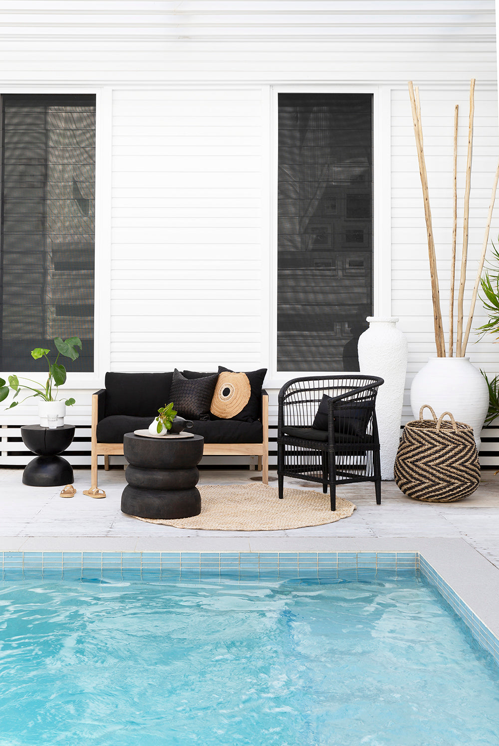 A stylish outdoor lounge setting featuring a black cushioned sofa with a natural wood frame, a black rattan armchair, and textured décor, set beside a serene poolside backdrop.
