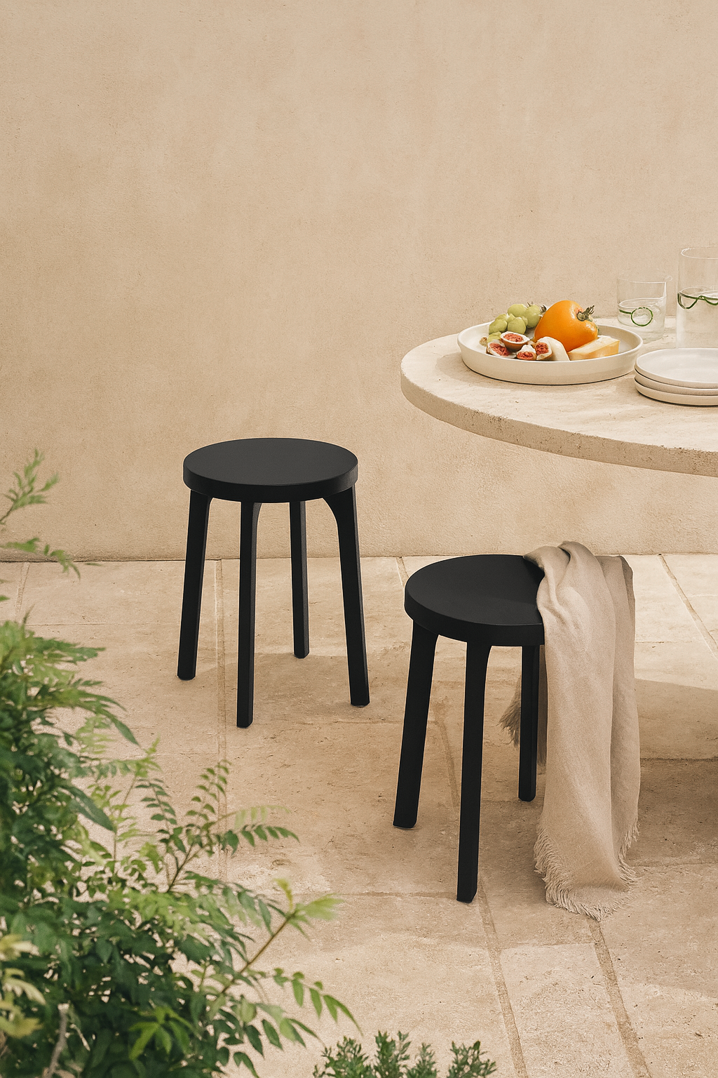 Pair of black outdoor stools with round seats styled in a sunlit courtyard next to a travertine table set with summer fruit and glassware