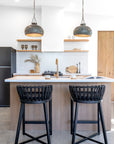 Black rattan bar stools styled at a timber kitchen island with white stone benchtop, brass tapware, and sculptural pendant lighting in a contemporary open-shelf kitchen