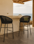Black rope outdoor bar stools with curved backs styled at a stone bar on a coastal balcony, overlooking the ocean with timber posts and a tiled floor