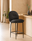 Black upholstered barstool with slim metal legs, styled beside a light wood counter in a neutral-toned room with natural light and minimal decor.