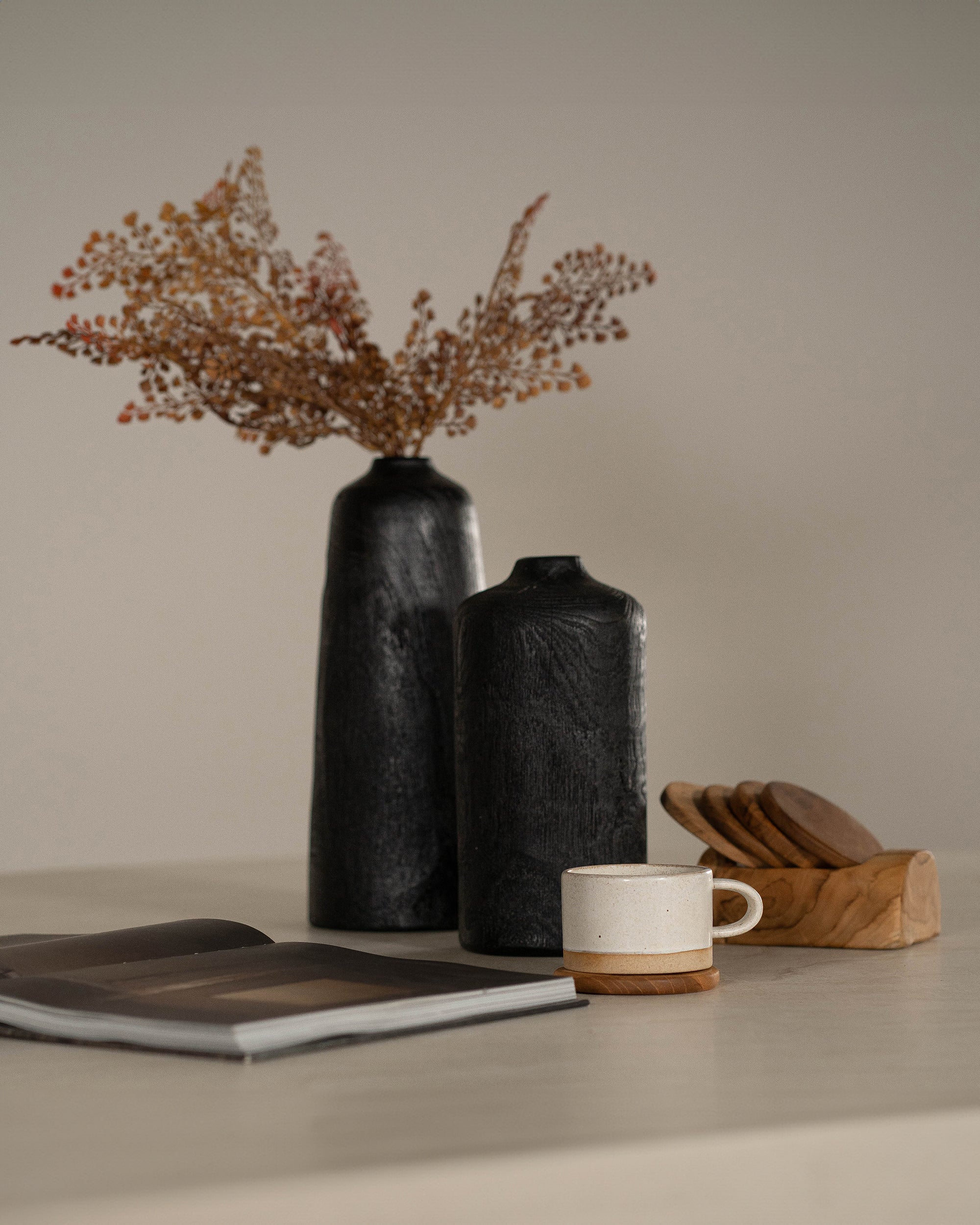 A moody and sophisticated interior setting featuring two hand-carved black wooden vases with dried foliage, a ceramic cup on a wooden coaster, and a stack of rustic timber coasters. A minimal open book adds a relaxed, curated touch to the scene
