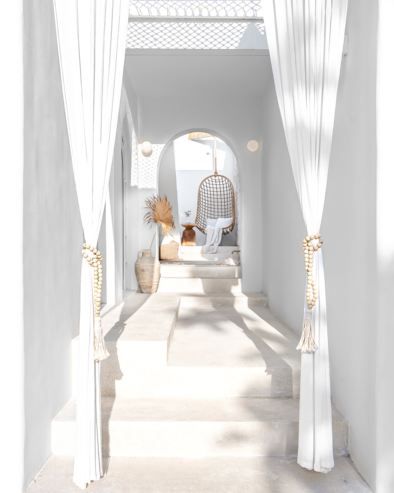 A serene bohemian-style setting featuring a natural rattan hanging chair framed by an elegant white archway. Sunlight filters through sheer white curtains, casting soft shadows on the stone steps, with decorative accents like dried palms, vases, and textured furnishings adding warmth