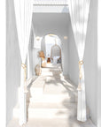 A serene bohemian-style setting featuring a natural rattan hanging chair framed by an elegant white archway. Sunlight filters through sheer white curtains, casting soft shadows on the stone steps, with decorative accents like dried palms, vases, and textured furnishings adding warmth