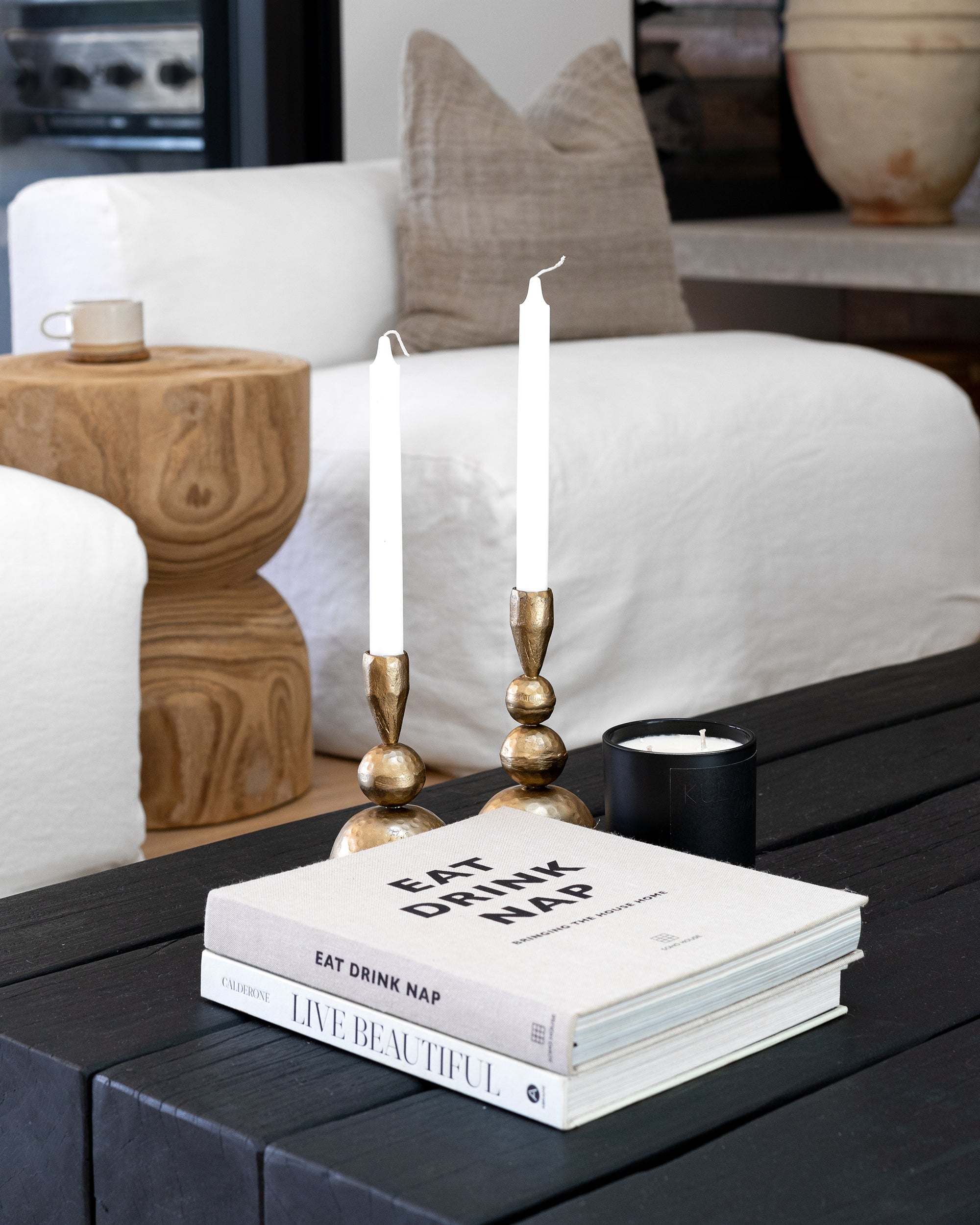 A refined coffee table vignette featuring KULALA Living’s brass candlestick holders with elegant tapered candles, paired with design books and a black minimalist candle. The setting is styled against a white linen sofa, a sculptural wooden side table, and neutral-toned accents, embodying a serene and sophisticated living space