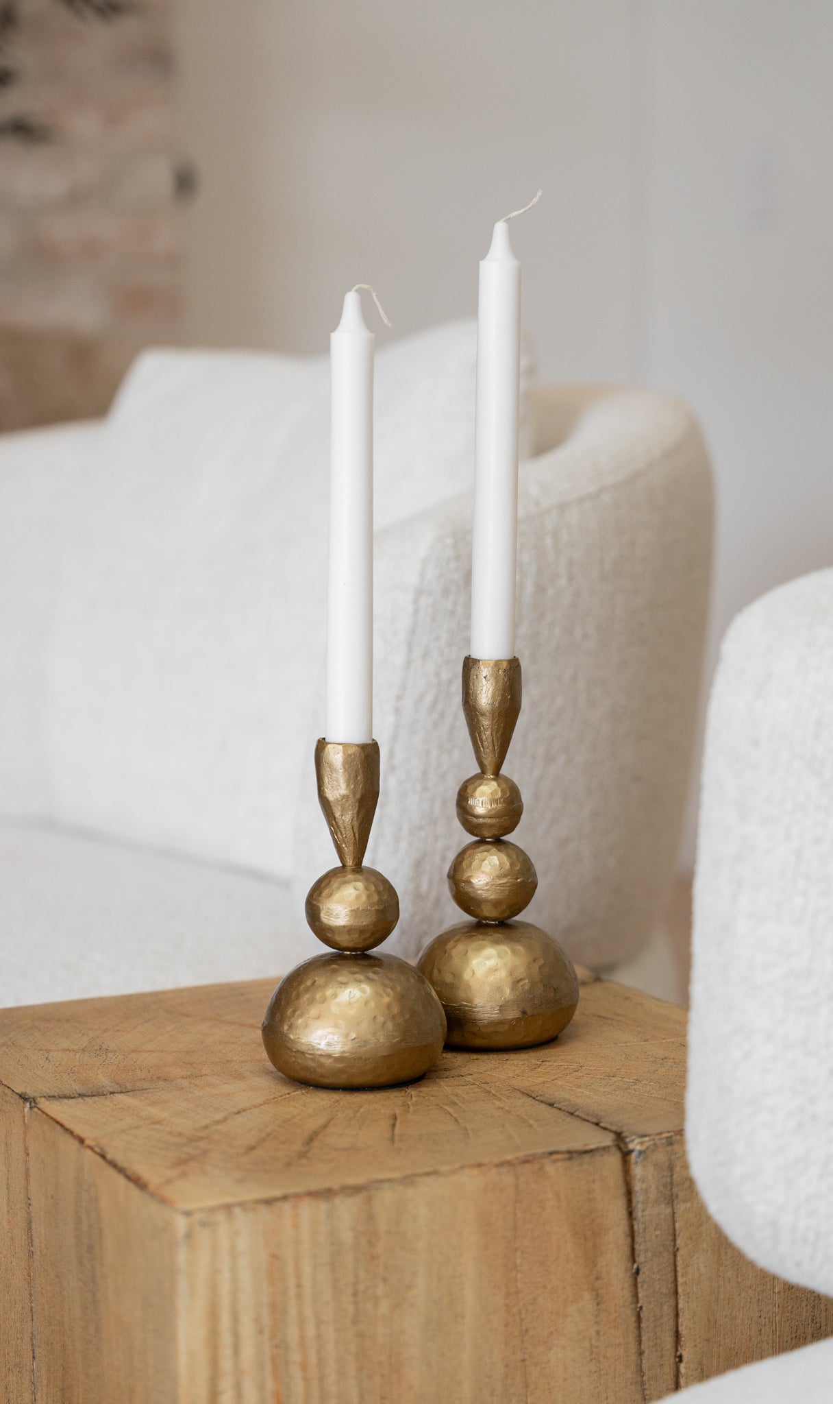 A pair of handcrafted brass candlesticks with white taper candles, elegantly styled on a solid timber side table. The warm brass tones contrast beautifully against the soft boucle chairs in the background, adding a touch of sophistication and warmth to the space