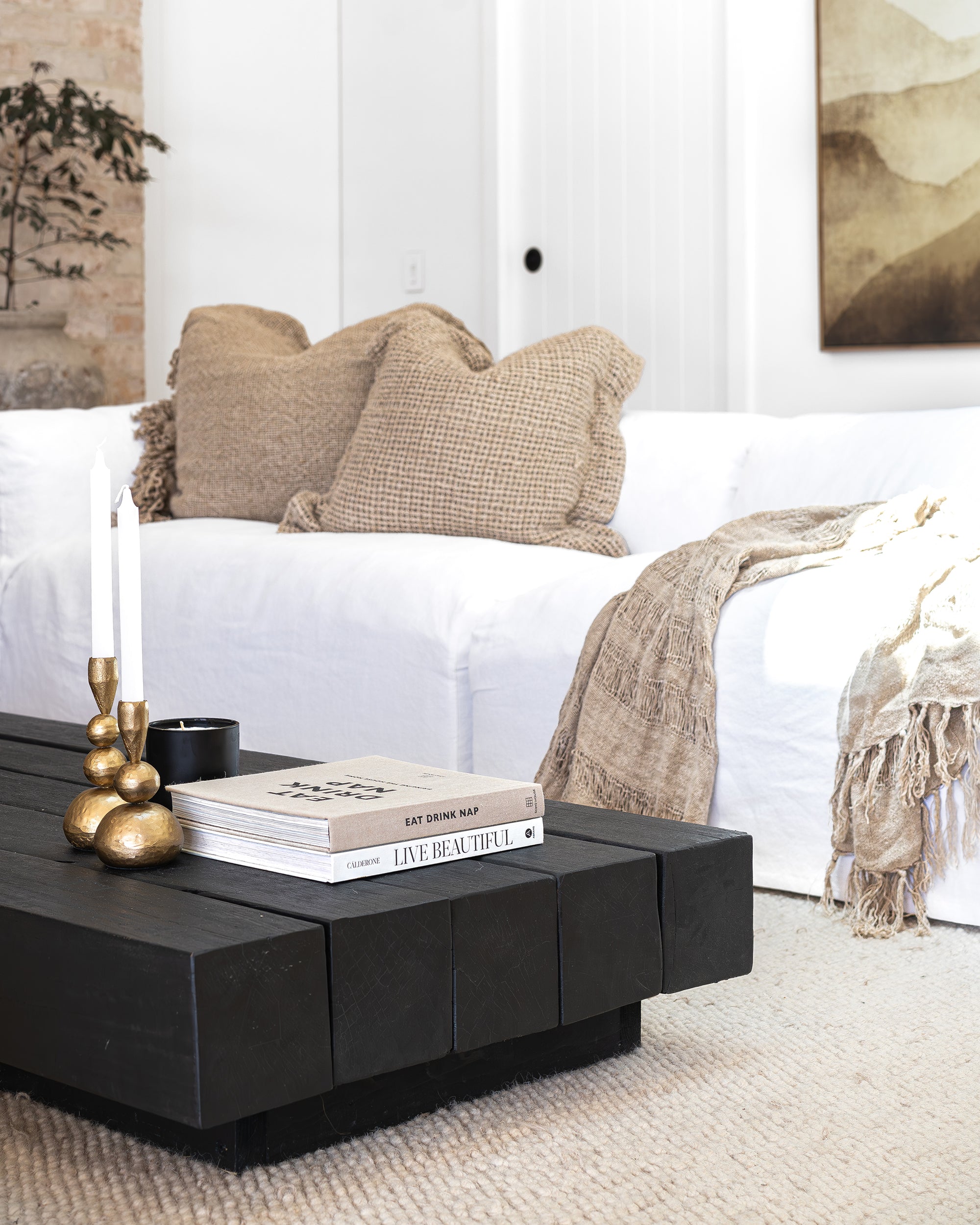 A beautifully styled modern living room featuring KULALA Living’s brass candlestick holders on a black timber coffee table. Neutral tones, textured cushions, and a soft woven throw create a warm and inviting aesthetic, complemented by curated books and a sleek black candle for a touch of sophistication