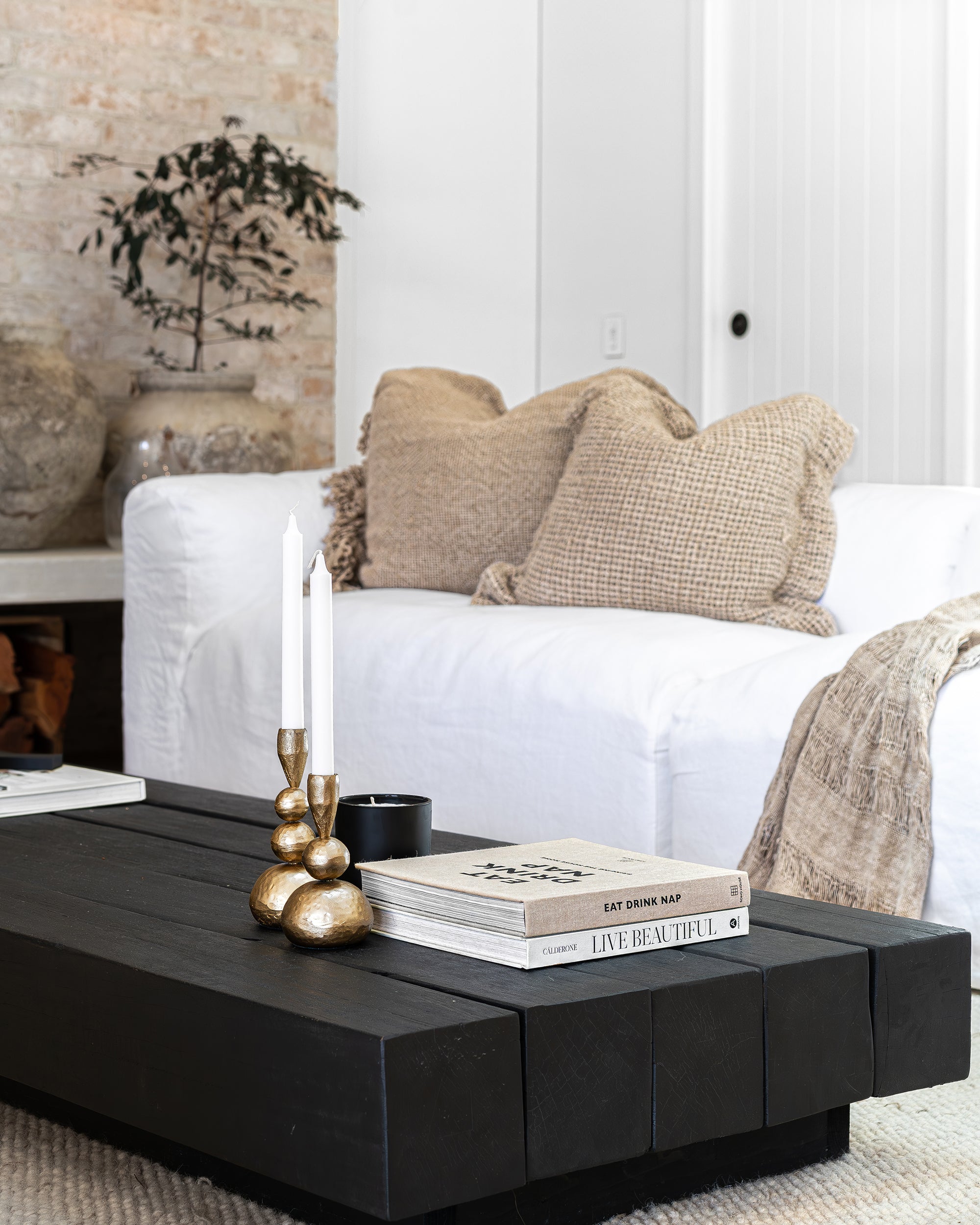 A beautifully styled living room showcasing KULALA Living’s brass candlestick holders on a black timber coffee table. The neutral palette, textured cushions, and soft woven throw create a serene and sophisticated atmosphere, complemented by curated books and a sleek black candle