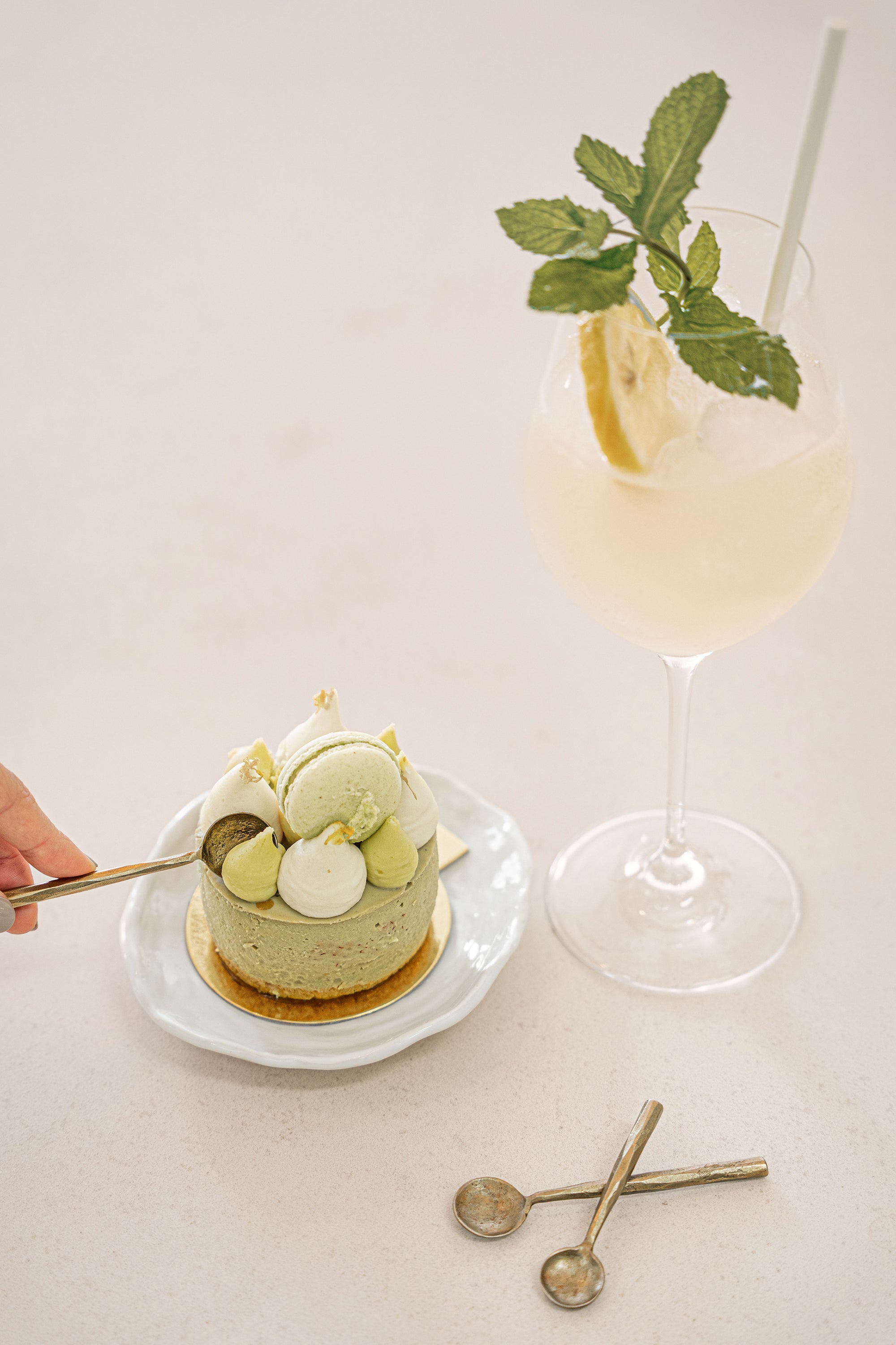 A luxurious dessert setting featuring KULALA Living brass dessert spoons, a matcha cheesecake topped with macarons, and a citrus-infused cocktail. A hand is seen delicately scooping a bite, embodying the essence of slow living and refined indulgence