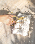 A hand holding vintage-style brass handle scissors, cutting twine around a beautifully wrapped linen bundle on a textured stone surface with natural sunlight and soft shadows