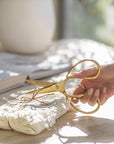 A hand holding vintage-style brass handle scissors, cutting twine around a beautifully wrapped linen bundle on a textured stone surface with natural sunlight and soft shadows
