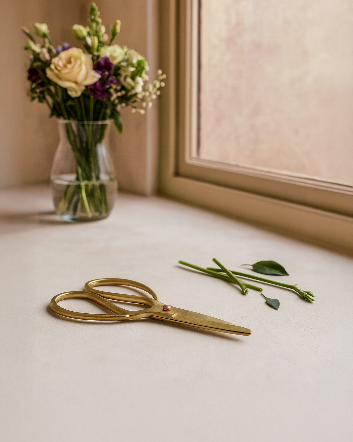 Brass Scissors styled beside freshly cut flower stems on a neutral surface near a window with a vase of blooms and soft natural light. 