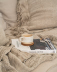 Close-up of a ceramic cup on the book 'This Is Home' resting on a textured knitted throw in a neutral-toned living space