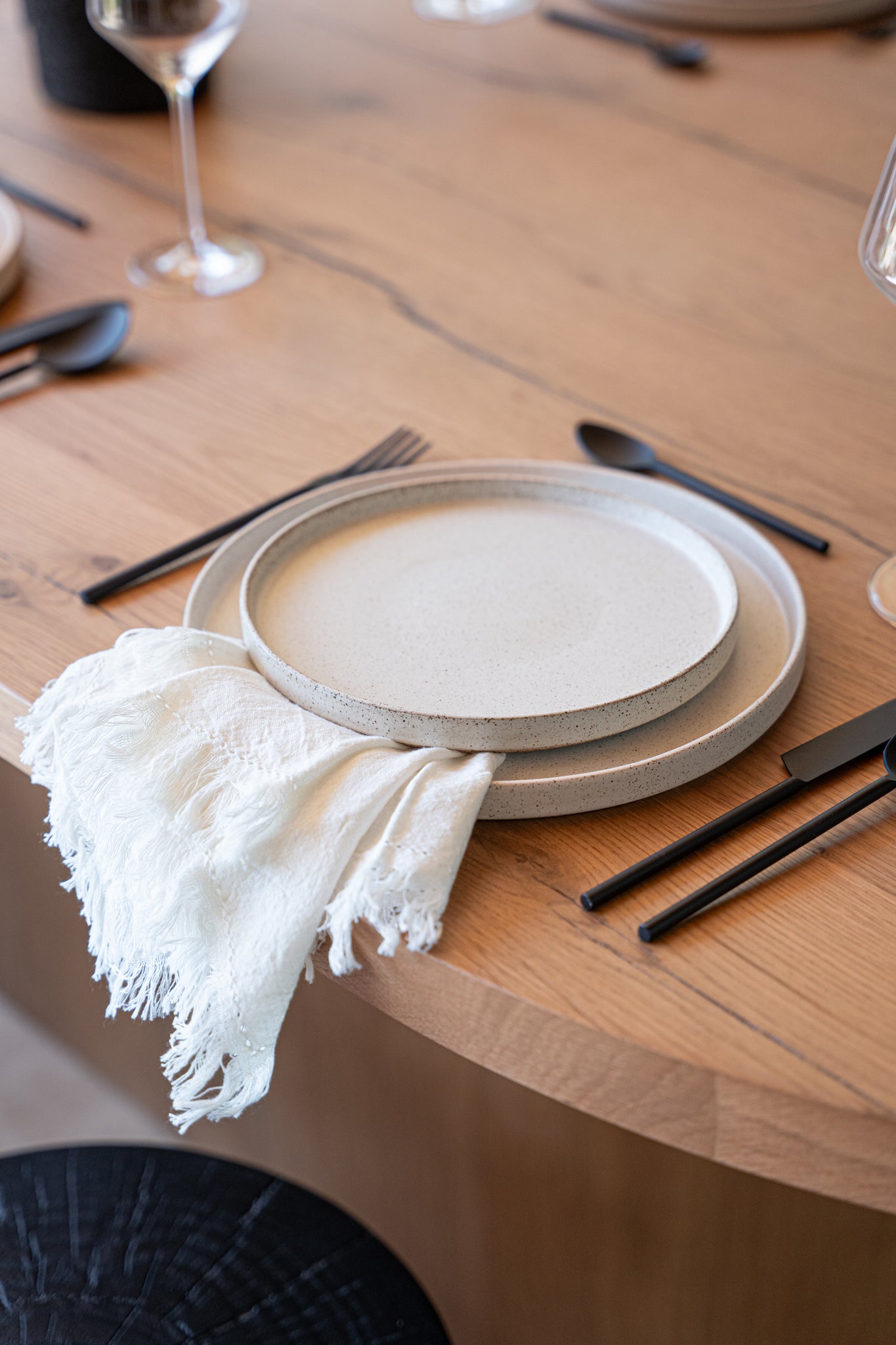 A modern dining setup featuring handcrafted ceramic dinner plates with a soft speckled finish, paired with matte black cutlery and a delicate fringed linen napkin, styled on a natural wood dining table.
