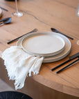 A modern dining setup featuring handcrafted ceramic dinner plates with a soft speckled finish, paired with matte black cutlery and a delicate fringed linen napkin, styled on a natural wood dining table.