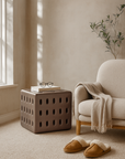 Contemporary cube-shaped side table in chocolate brown with cut-out detailing, styled in a cosy indoor setting beside a boucle armchair and soft neutral decor