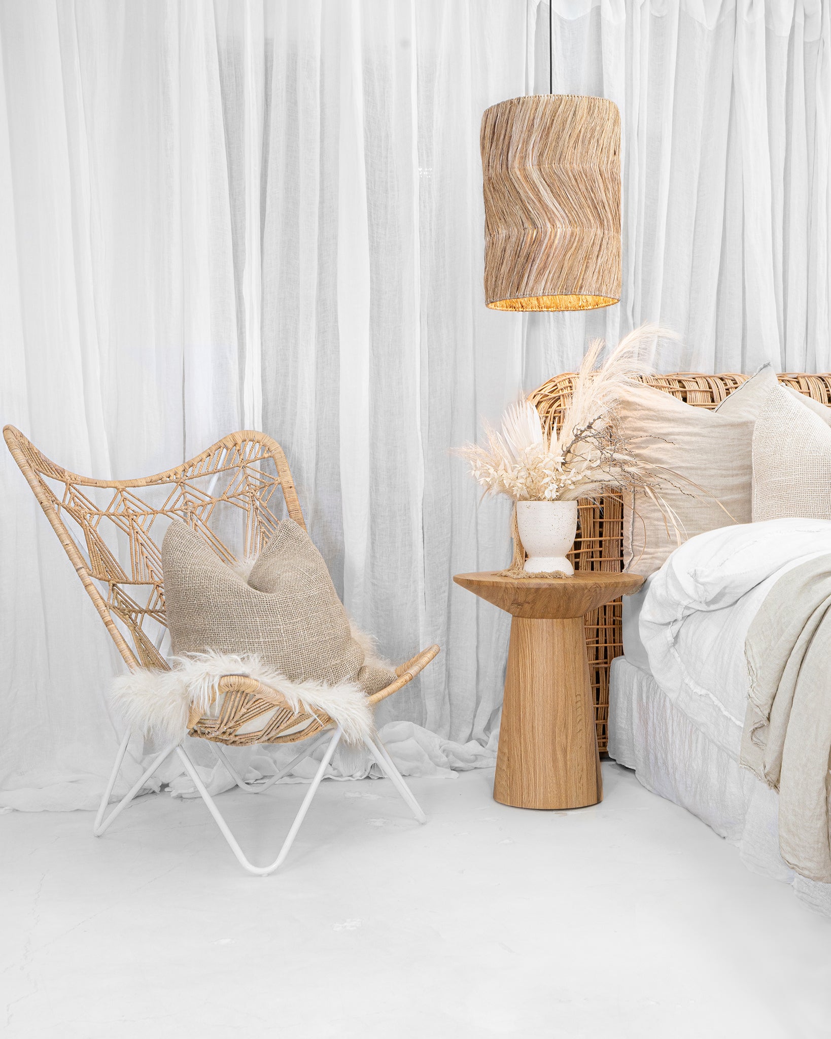 A serene coastal-inspired bedroom featuring a rattan bedhead, layered neutral linen cushions, a natural oak side table, and a woven pendant light, creating a relaxed and airy aesthetic