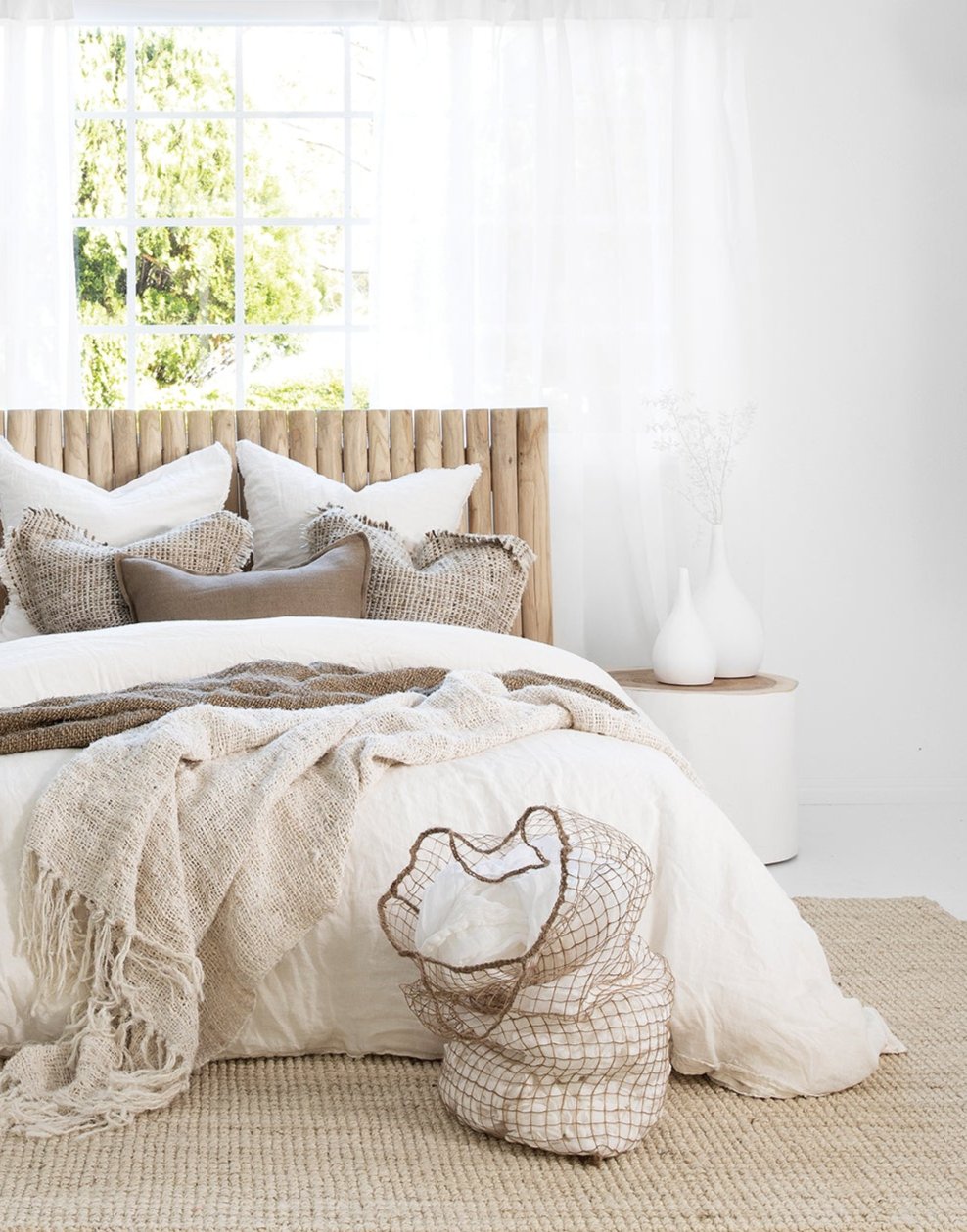 Tranquil coastal bedroom featuring a timber bedhead, layered neutral-toned bedding, woven cushions, and a soft jute rug, styled with white cement vases and a natural fibre laundry basket