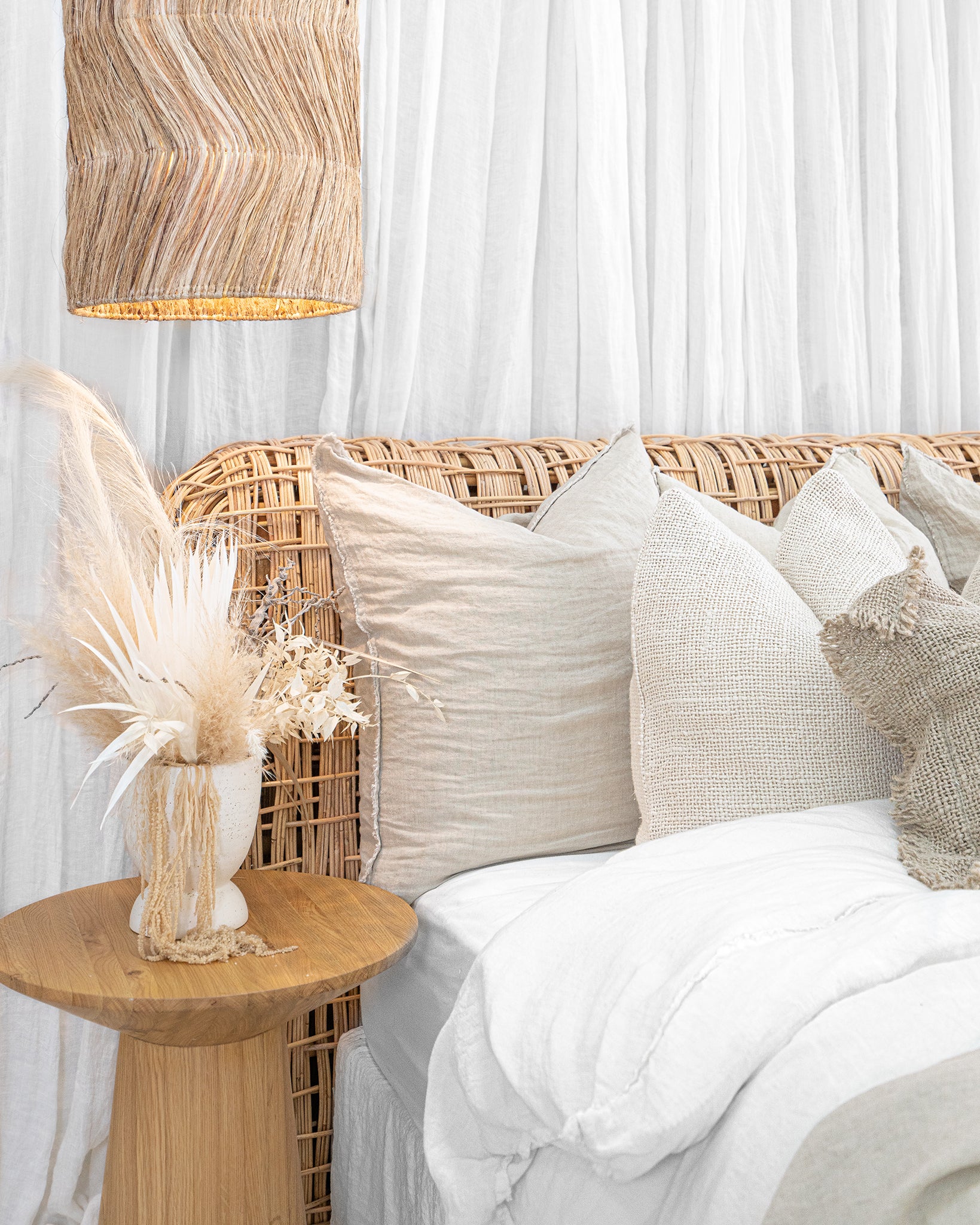 A serene coastal-inspired bedroom featuring a rattan bedhead, layered neutral linen cushions, a natural oak side table, and a woven pendant light, creating a relaxed and airy aesthetic