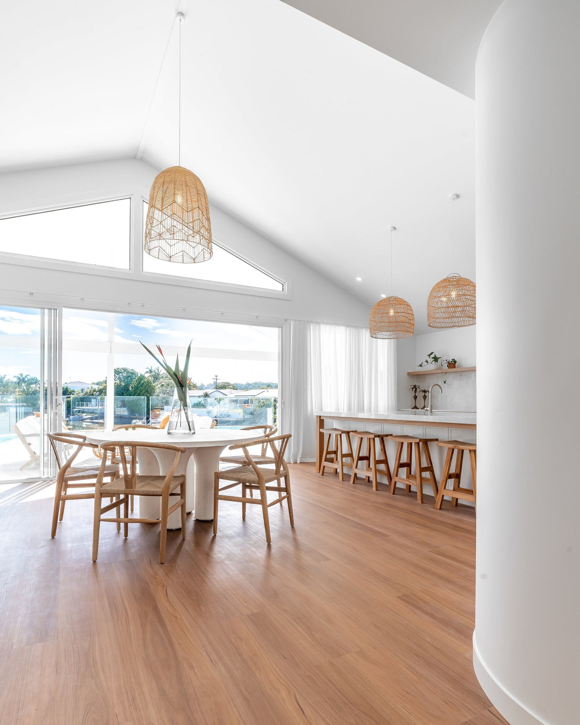A serene coastal dining space featuring natural wishbone dining chairs, a white sculptural dining table, and rattan pendant lights. The open-plan design flows seamlessly to an outdoor area with waterfront views, enhancing the bright and airy aesthetic