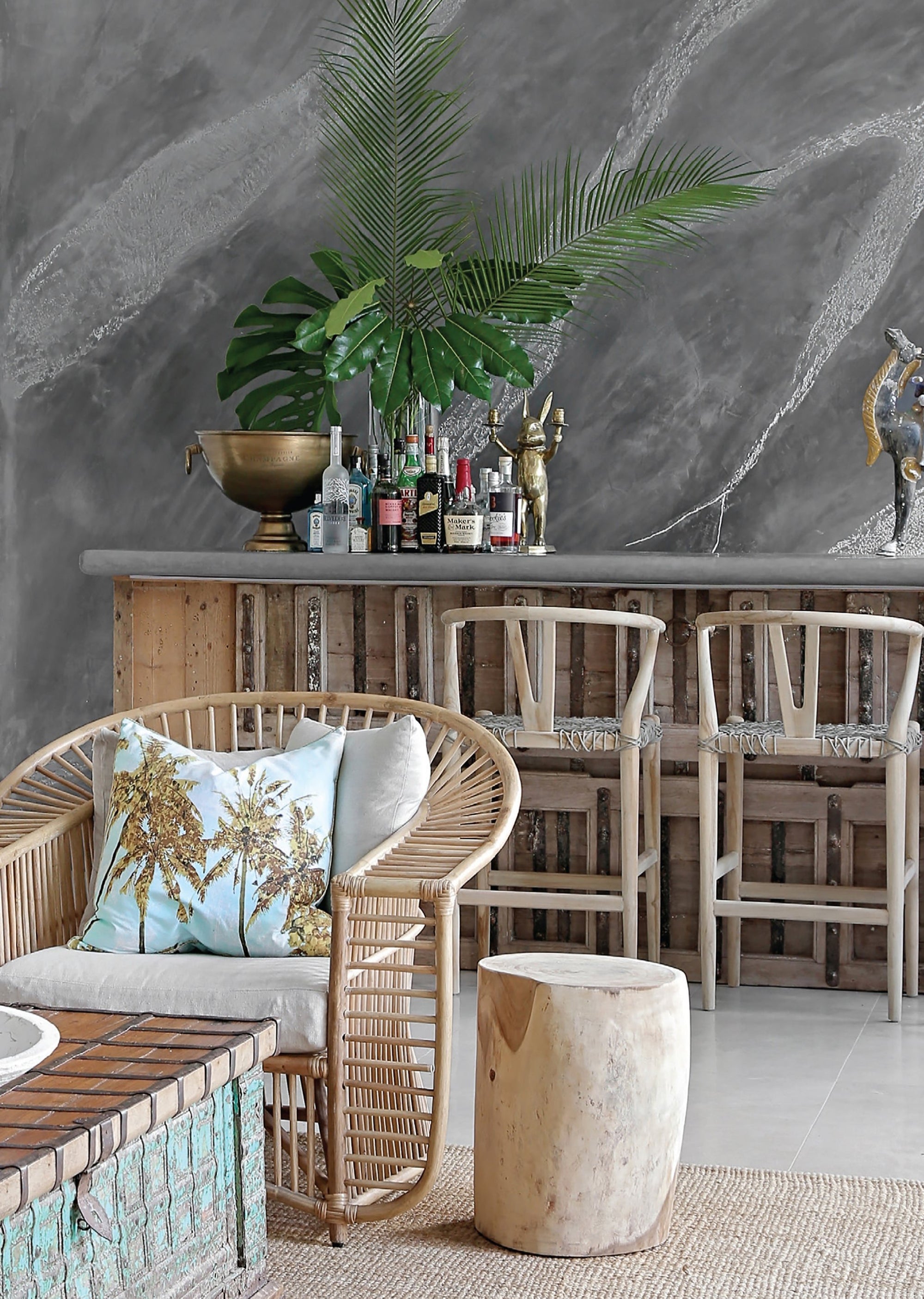 A stylish coastal bar setting featuring natural wood bar stools with woven seats, a rattan armchair with plush cushions, and a solid wood side table. The rustic bar area is complemented by tropical greenery and textured décor, creating a relaxed, resort-style ambiance