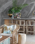 A stylish coastal bar setting featuring natural wood bar stools with woven seats, a rattan armchair with plush cushions, and a solid wood side table. The rustic bar area is complemented by tropical greenery and textured décor, creating a relaxed, resort-style ambiance