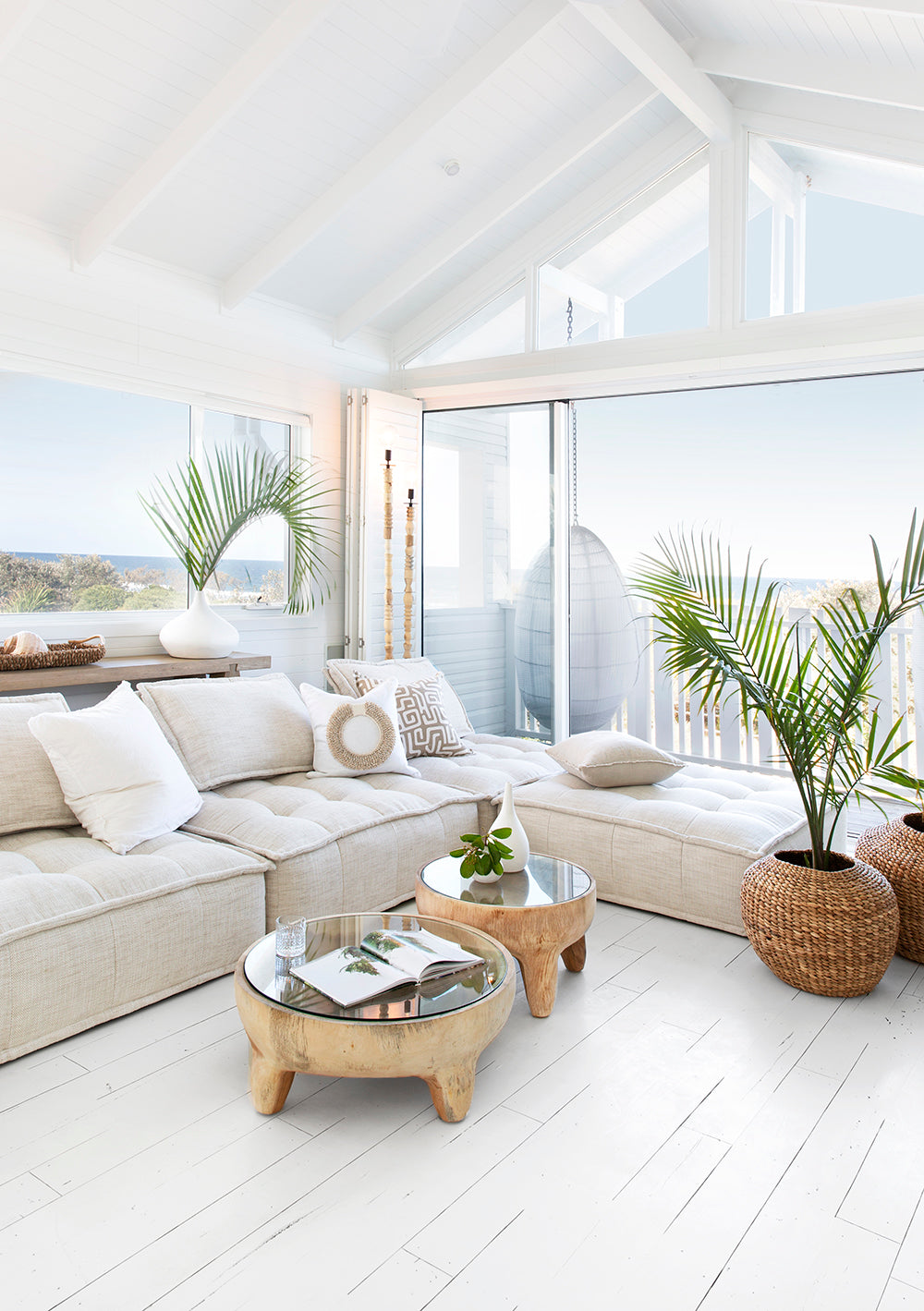 A serene coastal-inspired lounge featuring a plush white linen modular sofa, natural wood and glass-top coffee tables, and woven planters with lush greenery. Large windows offer breathtaking ocean views, creating a bright and airy retreat