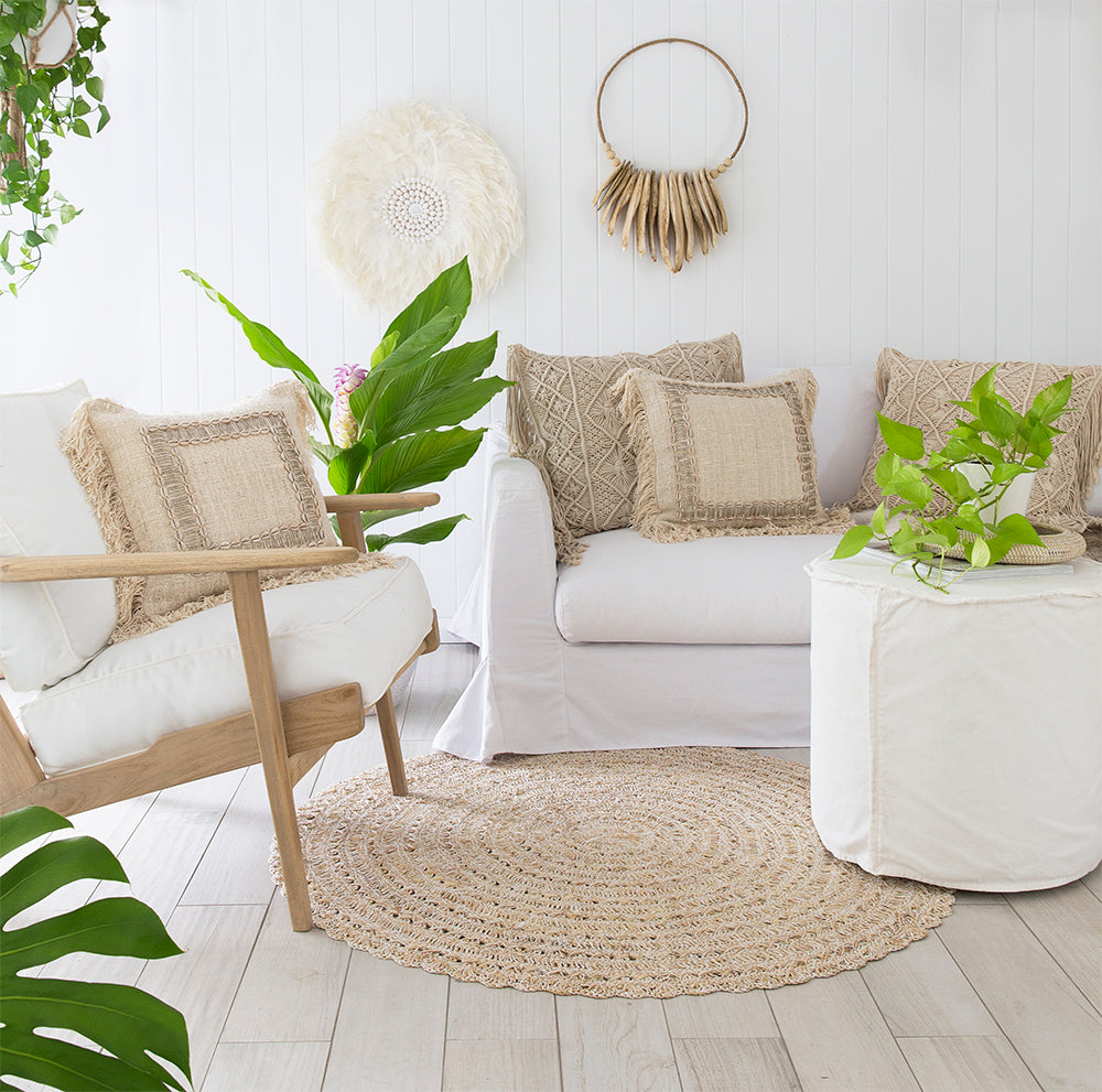 A serene coastal living space featuring a white slipcover sofa, natural woven cushions, and a linen slipcover ottoman. The setting is complemented by a rattan rug, wooden accents, and lush greenery, creating a relaxed and inviting atmosphere.