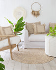 A serene coastal living space featuring a white slipcover sofa, natural woven cushions, and a linen slipcover ottoman. The setting is complemented by a rattan rug, wooden accents, and lush greenery, creating a relaxed and inviting atmosphere.