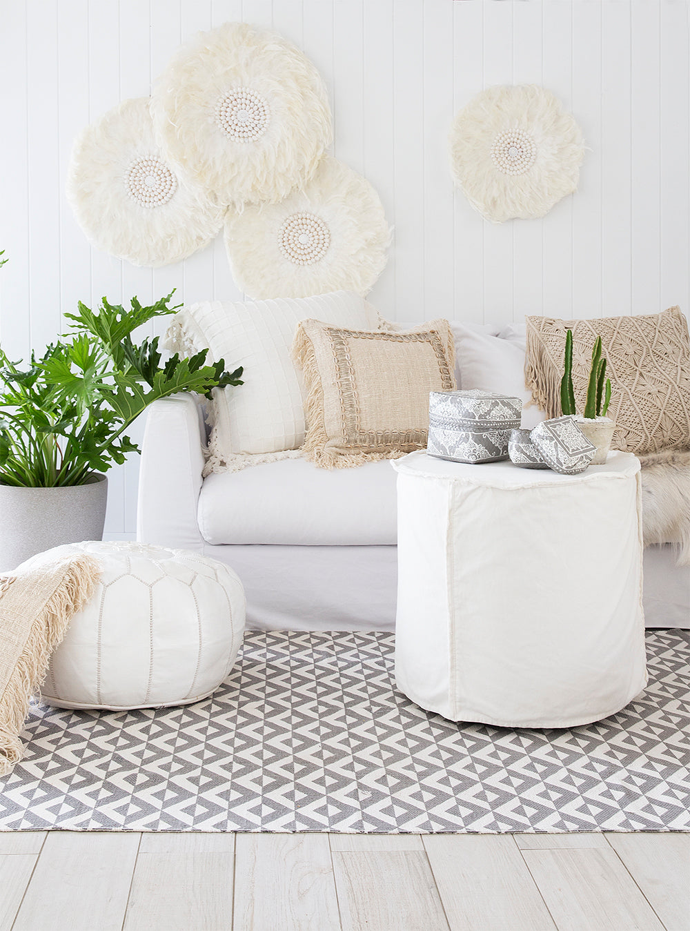 A serene coastal living space featuring a white slipcover sofa, natural woven cushions, and a linen slipcover ottoman. The setting is complemented by a rattan rug, wooden accents, and lush greenery, creating a relaxed and inviting atmosphere.