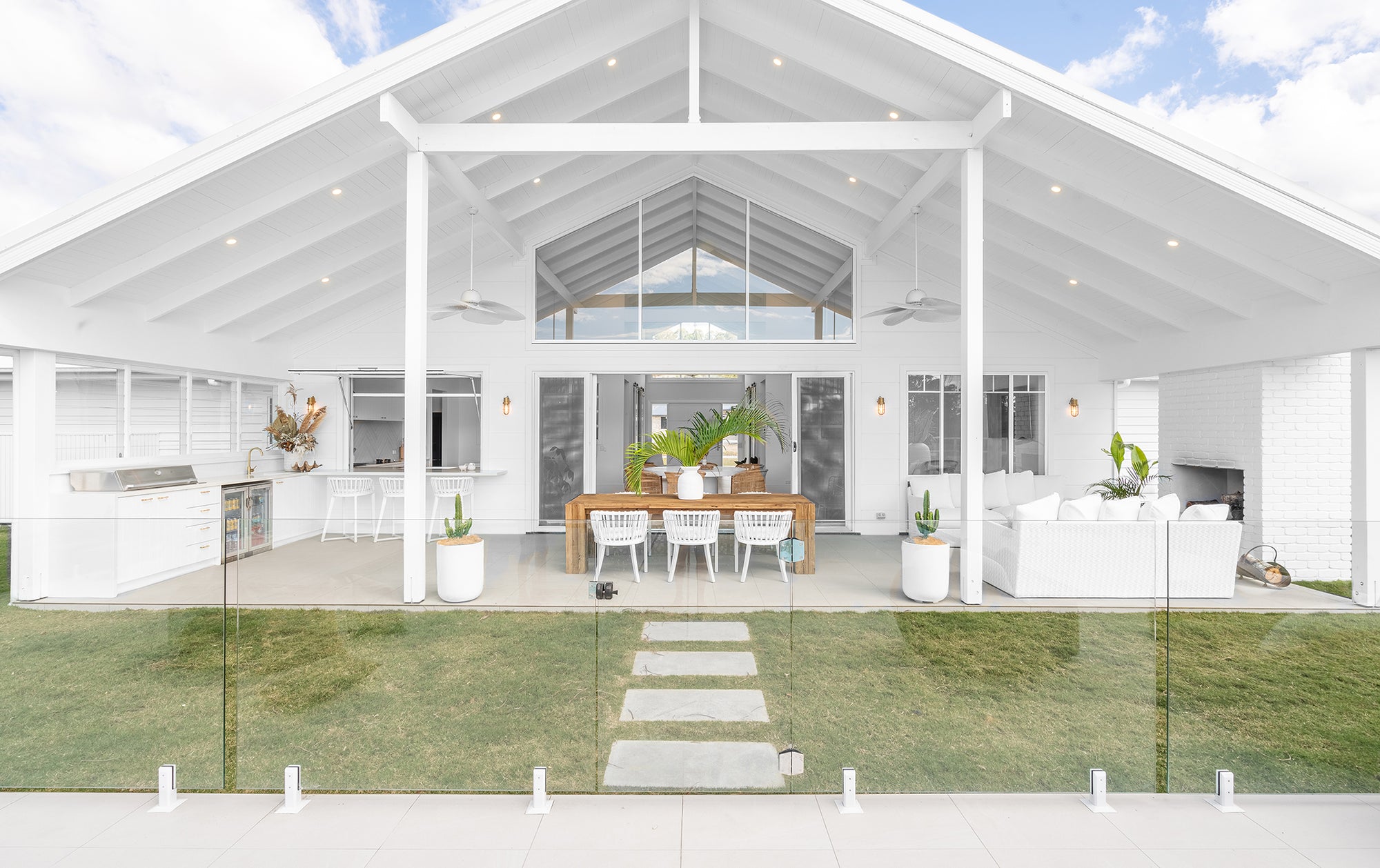 A beautifully designed coastal outdoor entertaining area featuring a spacious timber dining table with white Makena chairs, a built-in outdoor kitchen, and a cosy lounge setting. The gabled ceiling and open-plan design create a seamless connection between indoor and outdoor living