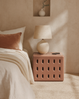 Concrete cube-shaped bedside table with modern cut-out design, styled in a serene bedroom with layered linen bedding and ceramic table lamp