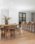Stylish open-plan dining and kitchen area featuring a natural oak dining table paired with woven rattan chairs and matching bar stools, creating a warm and inviting setting with layered textures and neutral tones.