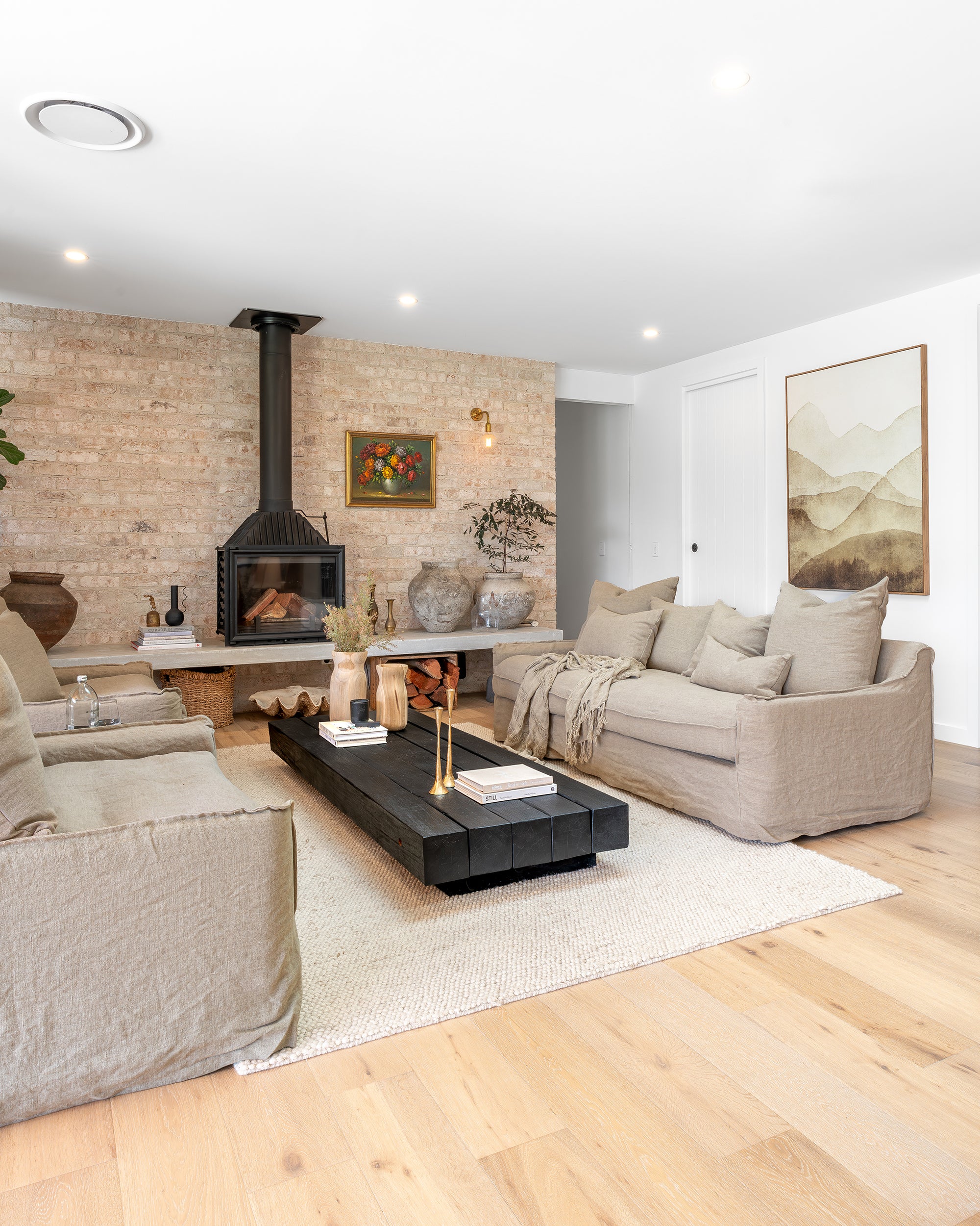 A stylish living space featuring natural linen sofas, a bold black timber coffee table, and a rustic brick fireplace. The neutral palette is complemented by organic décor, textured ceramics, and soft lighting, creating a cosy and sophisticated ambiance